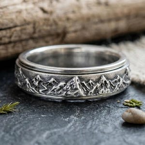 May include: A silver ring with a mountain range design. The ring features a detailed relief of mountain peaks around the band. The ring is made of a shiny, metallic material and is displayed on a dark surface.