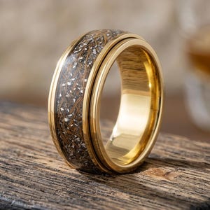 May include: A gold-colored ring with a dark, textured band. The band features a mix of brown and silver tones, creating a unique pattern. The ring is set against a wooden surface, with a blurred background.