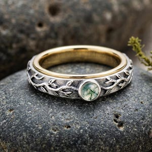 May include: A silver and gold ring with a green stone. The ring features a detailed vine and leaf design around the band. The central stone is a round, light green color. The ring is displayed on a gray stone.