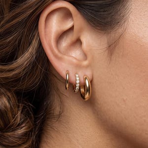Peut inclure: Trois créoles dorées portées sur un seul lobe d'oreille. La plus petite est lisse, celle du milieu est ornée d'une rangée de petites pierres claires, et la plus grande est un anneau épais, lisse et poli au profil arrondi et brillant.