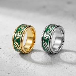 May include: Two rings with a green band featuring a white leaf design. One ring is gold-colored, and the other is silver-colored. The rings are set against a light gray background.
