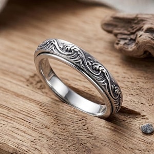 May include: A silver-toned metal ring featuring a continuous embossed pattern of stylized ocean waves around the outer band. The ring has a polished interior surface and a dark, oxidized finish within the recessed areas of the wave design.