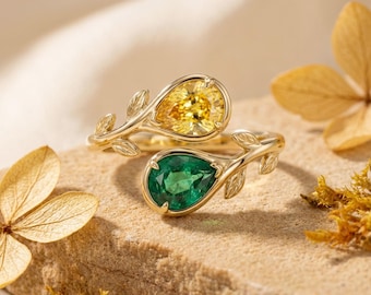 May include: Two gold rings with pear-shaped gemstones, one yellow and one green. The rings have a leaf design on the band. They are set on a textured, light brown surface, with dried flowers in the background. The gemstones are in a pear shape.