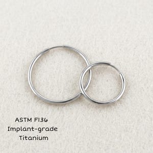 May include: Two silver-colored hoop earrings of different sizes made from ASTM F136 implant-grade titanium. The circular metal rings have a smooth, polished finish and are positioned side by side on a textured off-white surface.