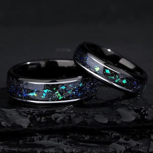 May include: Two black rings with a band of sparkling blue, green, and teal fragments. The rings have a polished black finish with a thin silver band. The rings are set against a dark background.