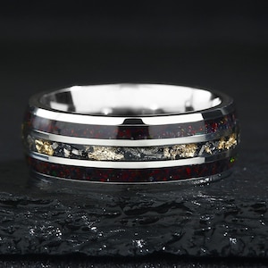 May include: A silver ring with a wide band. The ring features a dark red and black inlay with gold flecks. The ring has a polished, reflective surface and sits on a dark, textured surface. The ring's design is modern and eye-catching.