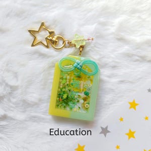 Puede incluir: Un llavero de resina verde y amarillo con un lazo y varios objetos pequeños en su interior. El llavero está unido a un llavero de estrella dorada. La palabra "Educación" está escrita debajo del llavero.