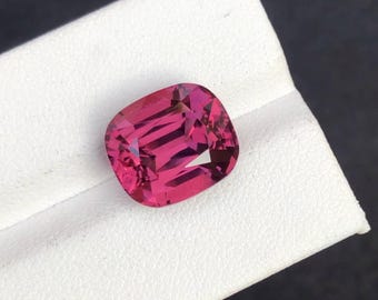 May include: A cushion-cut, vibrant pink gemstone with internal facets. The gem is set against a white surface, with a dark background. The gemstone's color ranges from deep magenta to lighter pink hues, showcasing its clarity and brilliance.
