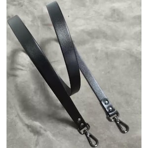 Può includere: Una tracolla per borsa in pelle nera con chiusure in metallo color argento. Il cinturino è sottile e flessibile, progettato per essere attaccato a una borsa o borsa. Le chiusure sono di colore argento lucido.