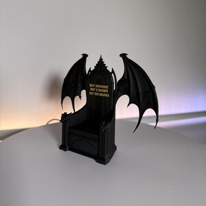 May include: A black miniature chair featuring large, detailed bat-like wings on either side of the backrest. The backrest displays the gold-coloured text "MY HOUSE MY CHAIR MY WOMAN" in a bold, capitalised font. The structure has a pointed top.