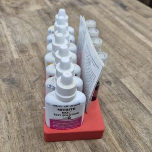May include: A red plastic rack holding multiple white plastic dropper bottles and a small glass vial. The front bottle is labeled Nitrite NO2 Test Solution with a pink accent. A folded white instruction booklet is tucked into the rack.