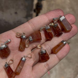 Pode incluir: Uma coleção de nove pequenos pingentes de cristal facetado na cor âmbar, cada um preso com um passador de metal prateado. Os cristais são translúcidos com tons variados de laranja e marrom, apresentando pontas e ferragens metálicas.