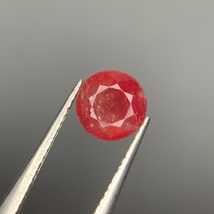 May include: A round, faceted red gemstone held by silver-toned metal tweezers. The stone features a brilliant cut with multiple triangular and kite-shaped facets that reflect light, displaying a deep, vibrant red color throughout its circular body.