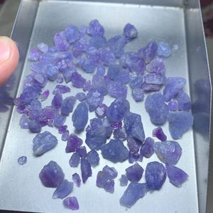 May include: A metal tray holds a scattering of small, rough-cut, blue-purple gemstones. The gemstones vary in size and shape, with a range of light and dark purple hues. The tray is held by a person's hand.