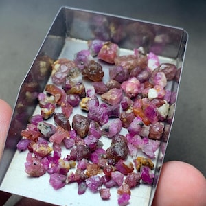 Puede incluir: Una colección de pequeñas piedras preciosas en bruto y sin pulir en tonos rosa intenso, magenta y marrón rojizo. Estas piedras irregulares y facetadas están contenidas en una bandeja rectangular de metal plateado con base plana.