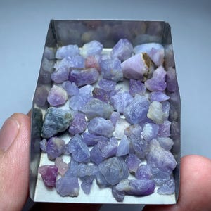 May include: A collection of small, raw, unpolished gemstones in shades of light purple, lavender, and soft blue. These irregular, translucent mineral fragments are contained within a rectangular, open-topped metal tray with a polished finish.