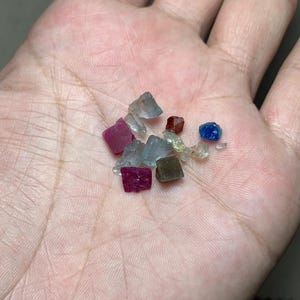 May include: A collection of small, colorful gemstones, including ruby red, sapphire blue, and clear crystals, are held in an open hand. The gemstones vary in shape and size, with a rough, natural appearance.