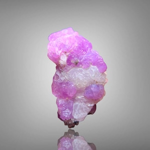 Puede incluir: Una gema en bruto sin cortar con un vibrante tono rosa, que exhibe una estructura cristalina. La piedra tiene una superficie rugosa y texturizada y una forma natural y orgánica. La gema se encuentra sobre un fondo neutro, con un reflejo visible debajo.