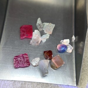 Op de afbeelding: Een verzameling ruwe edelstenen in verschillende kleuren, waaronder rood, roze, bruin en blauw, gepresenteerd in een kleine, zilveren bak. De edelstenen zijn onregelmatig van vorm en lijken ongeslepen, waardoor hun natuurlijke texturen en tinten zichtbaar zijn.