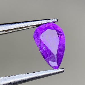 May include: A pear-shaped, faceted, vibrant purple gemstone held by silver tweezers. The gem's facets reflect light, showcasing its clarity and color. The background is a soft gray, highlighting the gem's brilliance. This image is ideal for jewelry and gemstone listings.