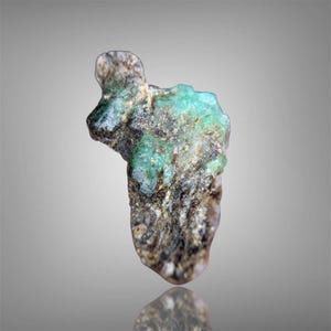 May include: A rough-cut emerald gemstone with a mix of green and brown hues. The natural stone has a textured surface and a reflective base, showcasing its raw beauty. The emerald is a precious gemstone.