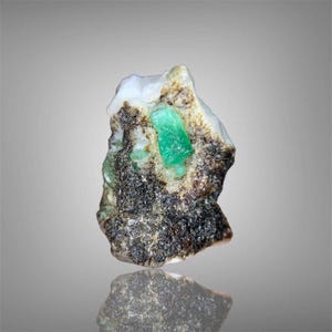 Pode incluir: Uma pedra preciosa de esmeralda bruta, apresentando um cristal verde vibrante embutido em uma matriz de rocha escura e salpicada e depósitos minerais brancos. A pedra é apresentada em um fundo neutro.