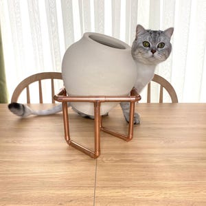 May include: A light beige ceramic vase with a wide opening, resting on a copper pipe stand. A gray cat with green eyes is peeking out from behind the vase. The vase and stand are on a wooden table.