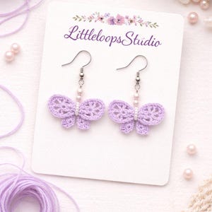 Puede incluir: Un par de pendientes de mariposa de ganchillo lavanda con ganchos plateados y detalles de perlas. Los pendientes se muestran en una tarjeta blanca con el texto "Littleloops Studio". El fondo incluye hilo lavanda y elementos decorativos.