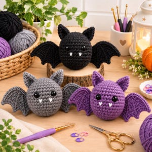 May include: Three crocheted bats in black, gray, and purple, each featuring round bodies, pointed ears, scalloped wings, black plastic eyes, and two small white felt fangs. The bats are arranged on a wooden surface with yarn and tools.