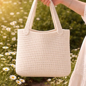 May include: Cream-coloured crochet tote bag with two sturdy handles. The bag has a rectangular shape and a textured surface, ideal for carrying items. The bag is held up in front of a field of daisies.