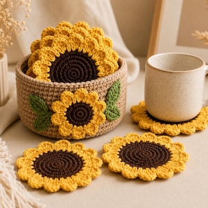 Pode incluir: Um conjunto de bases de copos de girassol de crochê artesanais. As bases amarelas com centros castanhos são exibidas em uma cesta tecida e ao lado de uma caneca. A cesta é decorada com girassóis e folhas verdes de crochê.