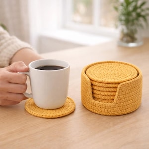 May include: A set of round, mustard yellow crocheted coasters with a matching circular holder. One coaster is placed under a white ceramic mug filled with dark coffee. The coasters feature a dense, textured stitch pattern in a vibrant yellow yarn.