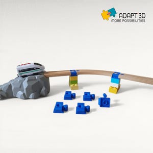 Puede incluir: Un juego de trenes de juguete con un tren blanco y verde sobre una formación rocosa gris, una vía de madera y conectores azules. La vía está sostenida por bloques de colores. Incluye el texto "ADAPT 3D MORE POSSIBILITIES".