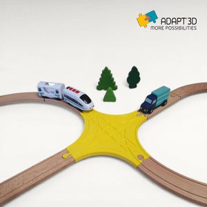 Op de afbeelding: Een houten speelgoedtreinset met een gele kruising. Een blauw-witte speelgoedtrein, een blauwe vrachtwagen en groene bomen staan op de baan. De tekst "ADAPT 3D MORE POSSIBILITIES" staat rechtsboven.