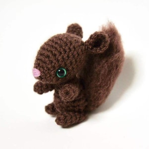 May include: A brown crocheted squirrel plush toy with green eyes and a pink nose. The squirrel has a fluffy tail.