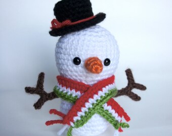 May include: A white crocheted snowman wearing a black top hat with a red band, a green and red scarf, and a brown twig for arms.