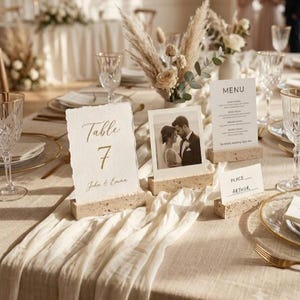 Op de afbeelding: Een bruiloftstafelsetting met een getextureerde papieren tafelnummerkaart met de tekst Table 7, een vierkante foto van een koppel, een gedrukt menu en een naamkaartje, allemaal geplaatst in rechthoekige standaards van travertijnsteen.