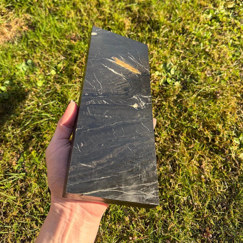 May include: A black rectangular stone with white veining and a gold-colored streak. The stone is held in a hand against a backdrop of green grass. The stone has a polished surface and a smooth texture.