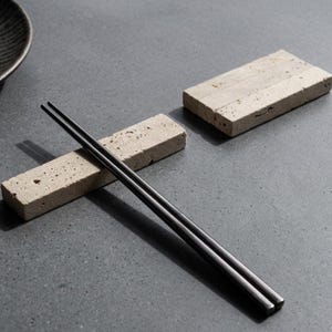 May include: A pair of sleek, black, rectangular chopsticks resting on a porous, beige, rectangular stone chopstick rest. A second, identical beige stone chopstick rest is positioned nearby, showcasing the natural, pitted texture of the stone.