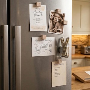 May include: A stainless steel refrigerator door featuring four square cork magnets holding a menu, a black and white photograph, a travel itinerary, and a grocery list. A small glass mason jar attached to the door contains several pens.