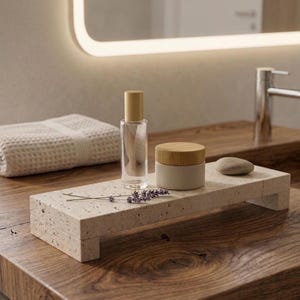 Op de afbeelding: Een rechthoekige beige gespikkelde stenen verhoger met vier korte pootjes. Op het oppervlak staan een doorzichtige glazen fles met een gouden dop, een cilindrische houder met een lichtbeige basis en houten deksel, en een takje gedroogde lavendel.
