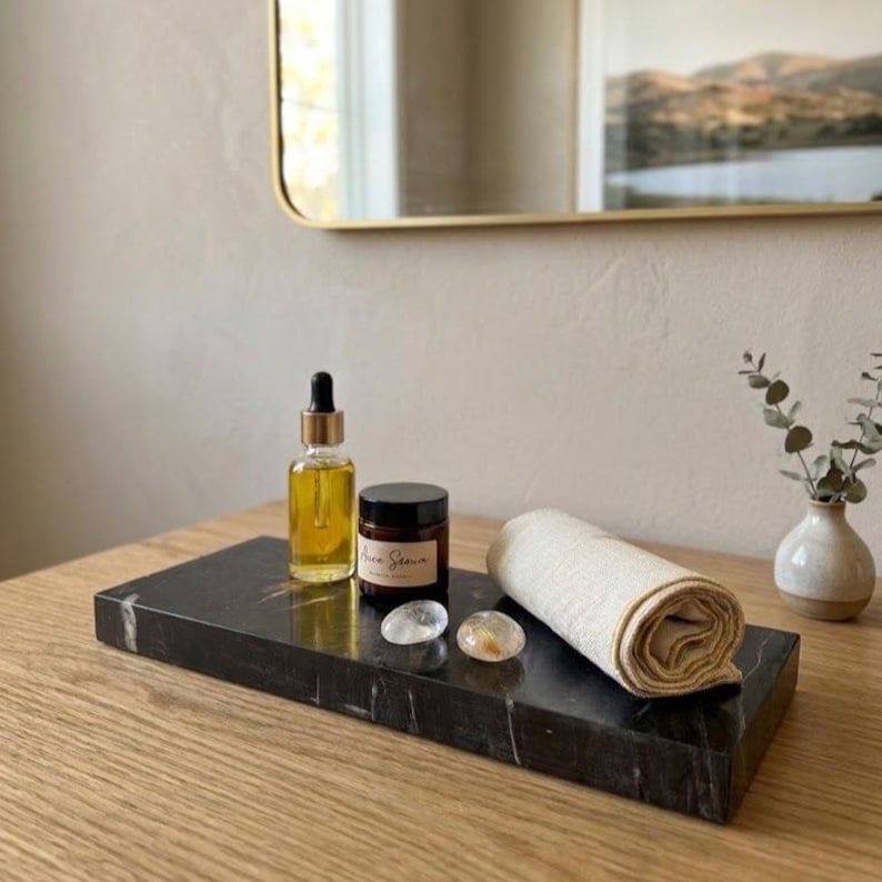 May include: A black marble tray holds skincare products: a dropper bottle of oil, a jar with a label, two clear stones, and a rolled-up beige cloth. A small vase with greenery is on the right.