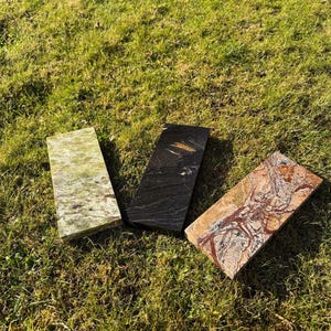 May include: Three rectangular stone slabs with unique patterns and colors are displayed on a grassy surface. One slab is green, another is black with gold veining, and the third is brown with red and white veining. The slabs are likely for decorative or construction purposes.