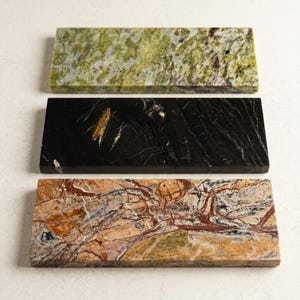 May include: Three rectangular stone slabs with unique patterns. The top slab is green with dark spots, the middle is black with gold veining, and the bottom is tan with red and brown veining. These decorative slabs are likely used for serving or display.