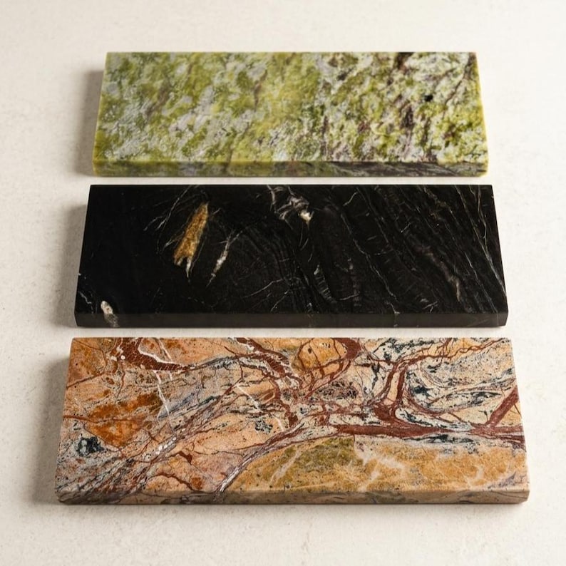 May include: Three rectangular stone slabs with unique patterns. The top slab is green with dark spots, the middle is black with gold veining, and the bottom is tan with red and brown veining. These decorative slabs are likely used for serving or display.