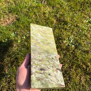 May include: A rectangular, polished stone with a mottled pattern of green, brown, and cream colors. The stone is held in a hand against a backdrop of green grass. The stone's surface has a natural, organic appearance.