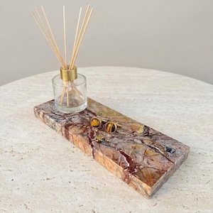 May include: A rectangular marble tray with a natural stone pattern in shades of brown, red, and beige. A small glass diffuser bottle with reeds and gold accents sits on the tray, along with a pair of gold cufflinks with tiger's eye stones.