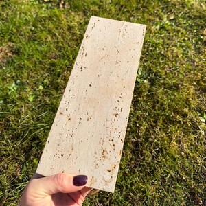 May include: A rectangular piece of beige travertine stone with a porous, textured surface. The stone features natural variations in color and small, irregular pits throughout its flat face, showcasing the characteristic appearance of this natural material.