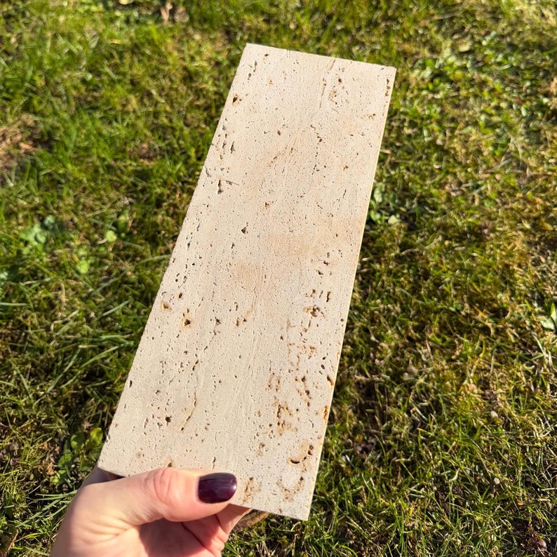 May include: A rectangular piece of beige travertine stone with a porous, textured surface. The stone features natural variations in color and small, irregular pits throughout its flat face, showcasing the characteristic appearance of this natural material.