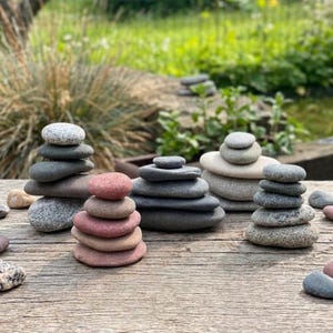 Puede incluir: Varias pilas de piedras lisas y planas de varios tamaños y colores, incluyendo gris, rosa y beige, están dispuestas sobre una superficie de madera envejecida. Las piedras están apiladas para crear pequeñas torres equilibradas, con un fondo verde borroso.